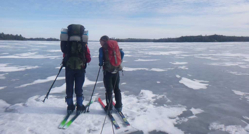 Two people wearing backpacks stand on cross country skis on a frozen lake. 